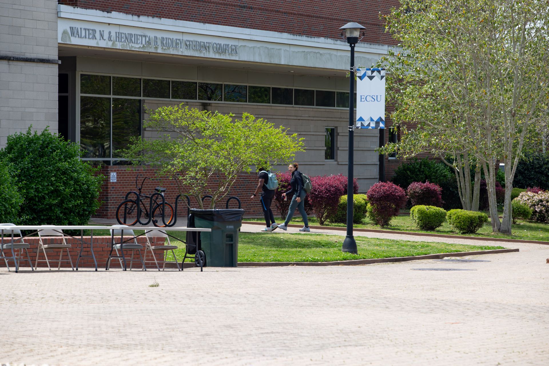 Students walk along a pathway outside the Walter N. & Henrietta B. Ridley Student Complex at ECSU. Trees, landscaping, bike racks, and an ECSU banner are visible in the campus courtyard.