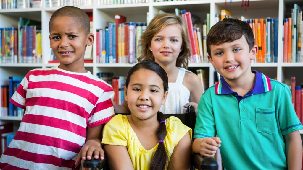 Four cheerful children smile together in a library classroom. One girl with long brown hair sits in a wheelchair. The children are posing in front of shelves filled with books. The sunny daytime photo would be great for projects about education, childhood, schools, and inclusion
