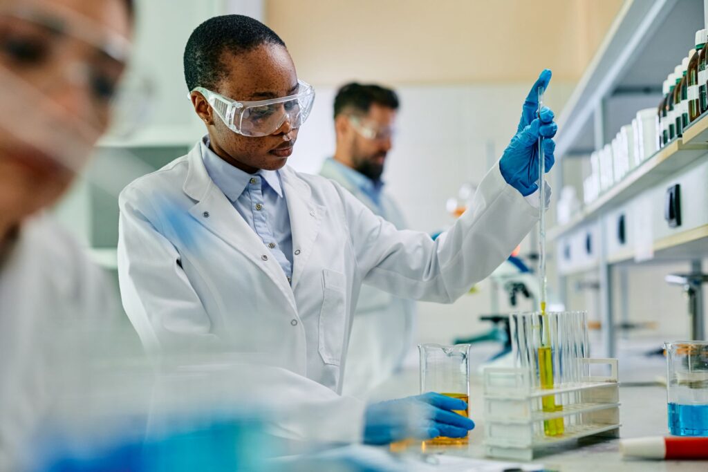 A woman in a lab coat and safety glasses is carefully using a pipette to transfer liquid into test tubes. 