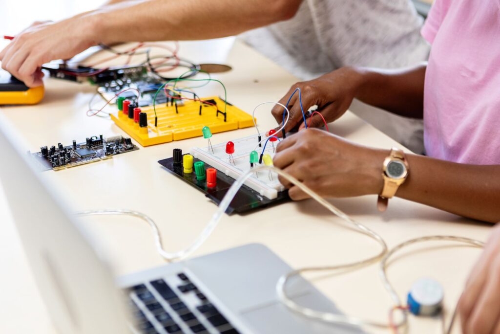 Two adults work together on an electronic project at a light-colored table.