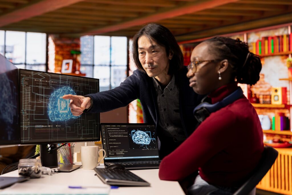 A man and a woman are working on computers with images of an artificial intelligence displayed on the monitors.