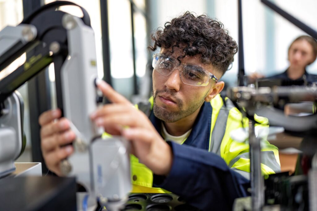 A young man with curly brown hair works with a large robotic arm in what appears to be a modern workplace.