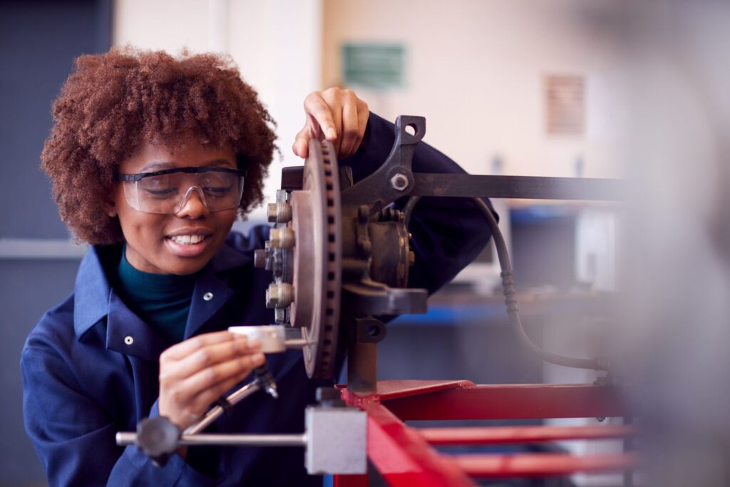 A young woman with curly brown hair wearing safety glasses smiles as she carefully examines a car part mounted on a red metal frame. 