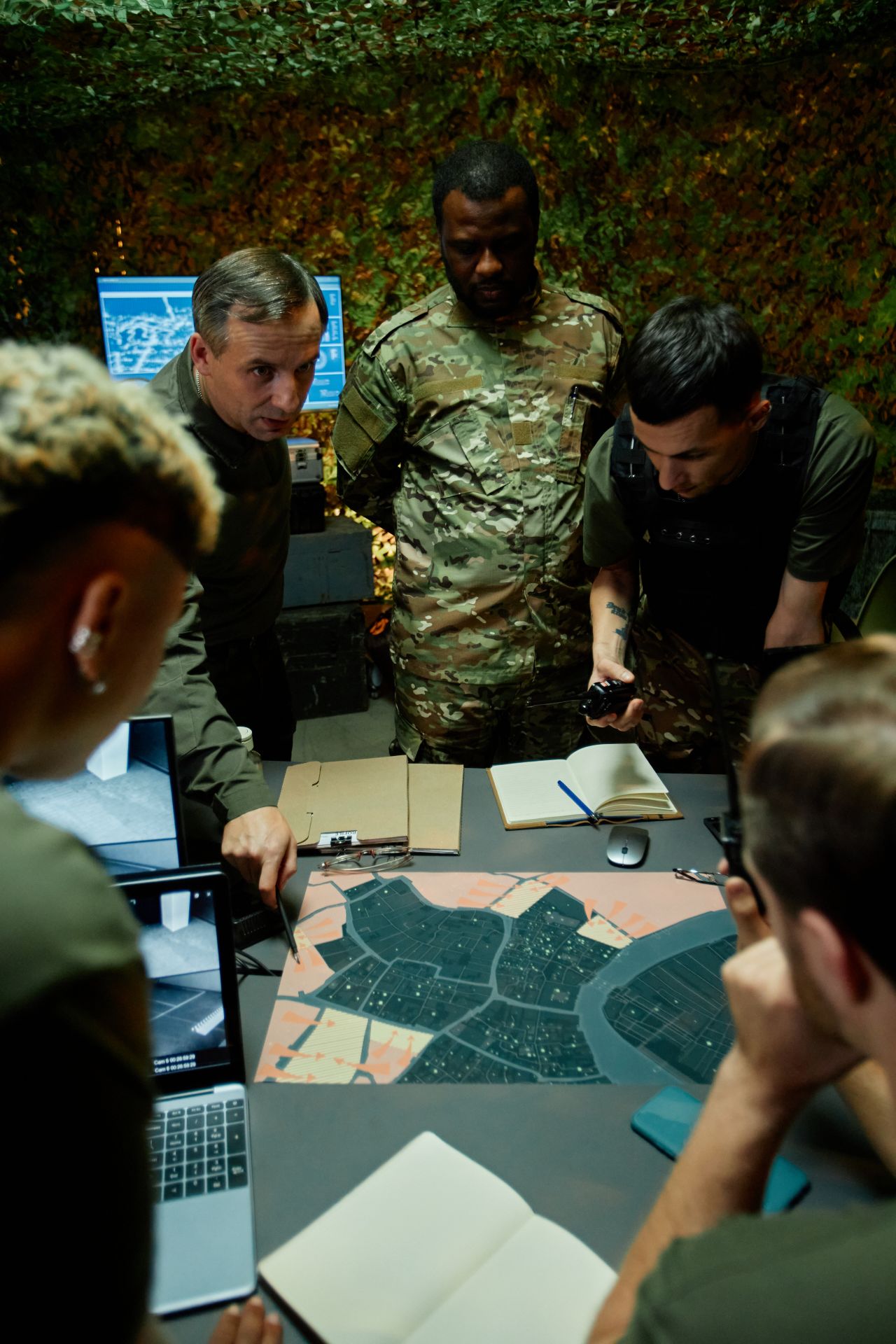 Six people gather around a table in what appears to be a camouflage-decorated office space.