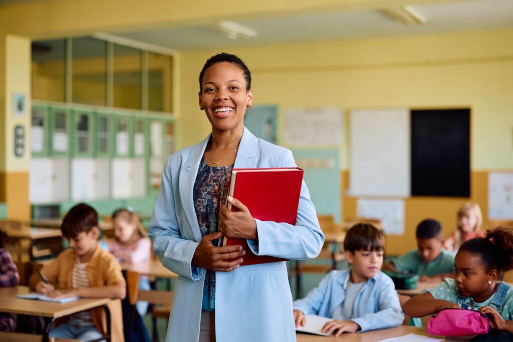 A teacher smiles broadly as she holds a stack of books in a classroom, students work at their desks in the background. 
