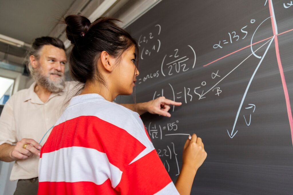 A young person with their hair up in a bun stands facing a chalkboard, writing math equations and diagrams.