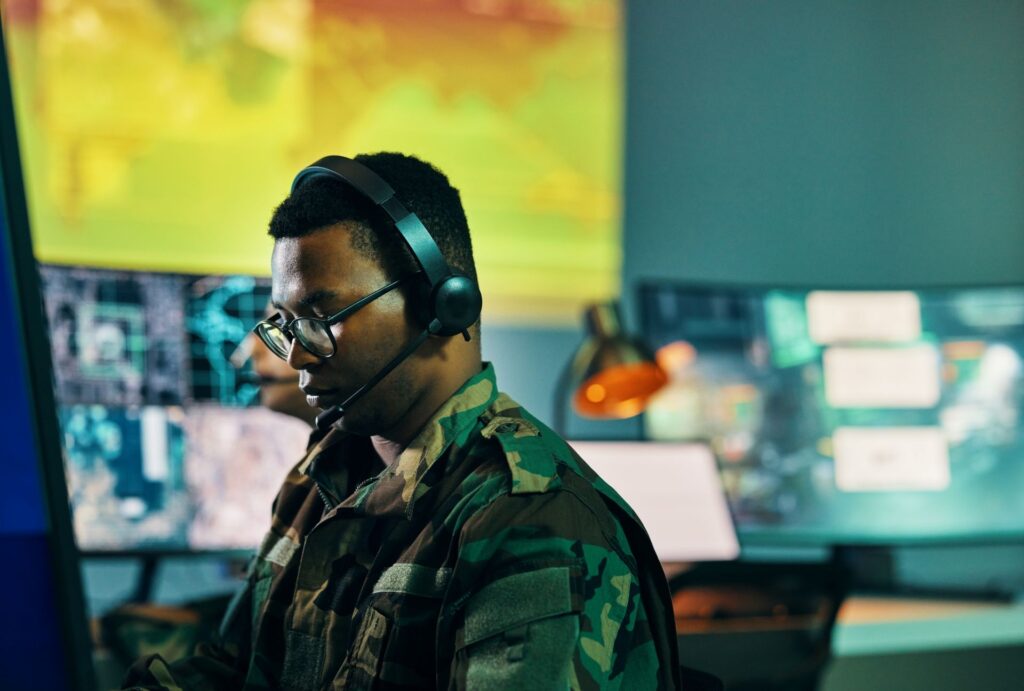 A man with curly dark hair wearing glasses and a camouflage jacket sits in front of multiple computer screens.