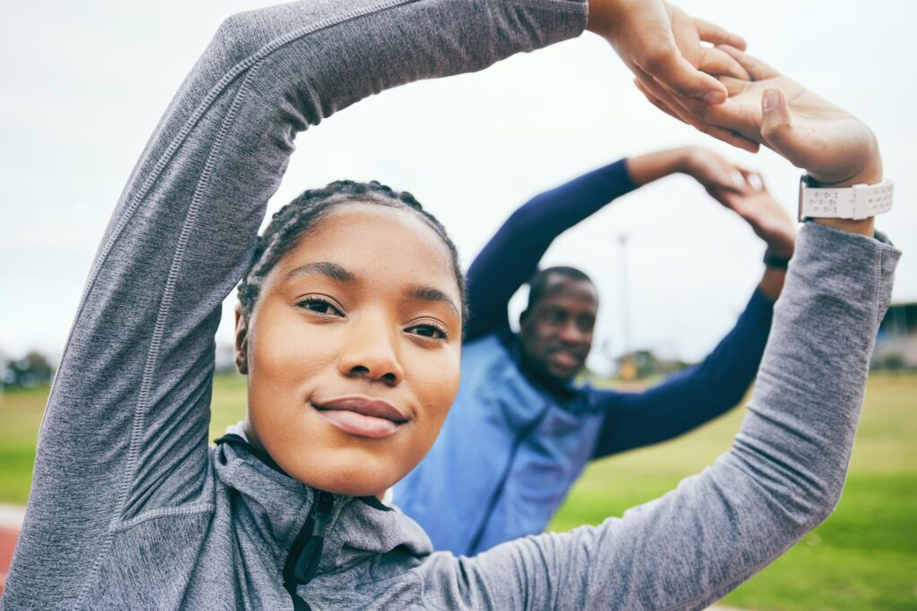 A young woman in a grey jacket smiles while stretching her arms overhead, creating a circle with her hands.