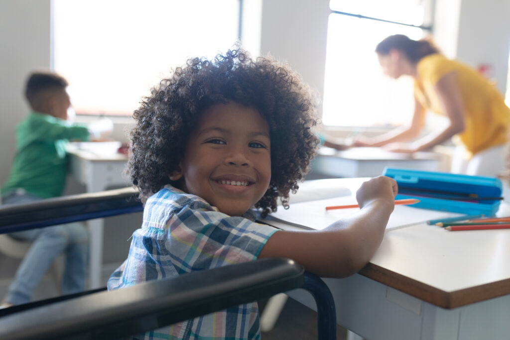 A smiling boy with curly hair is sitting at a school desk in a classroom. 
