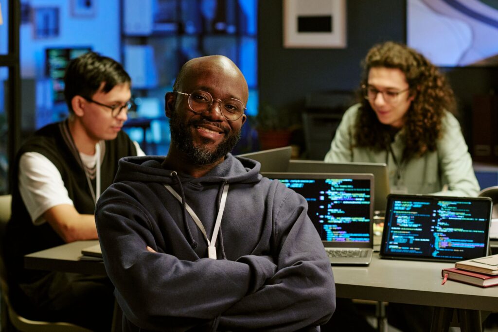 A smiling man with a beard, wearing glasses and a hoodie with arms crossed, poses in front of a table with laptops displaying lines of code, along with two coworkers.