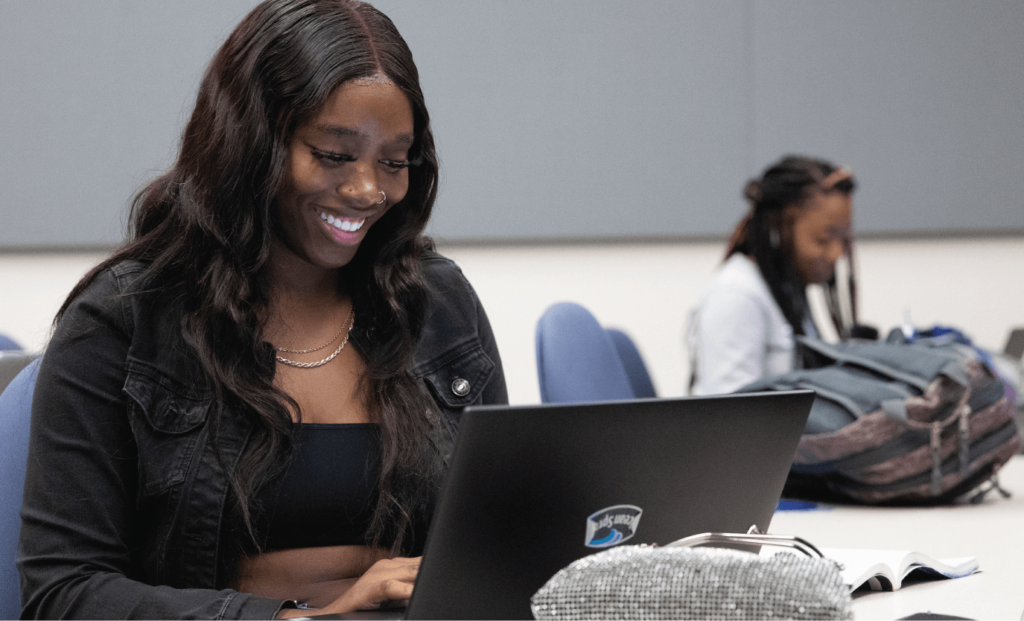 Student works on a laptop during class while another student studies in the background.