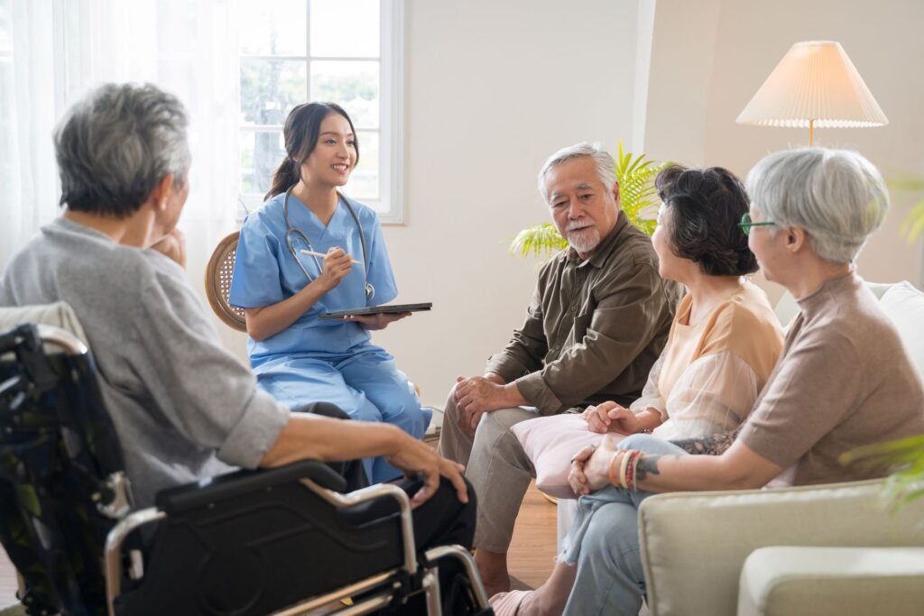 A young adult female nurse in blue scrubs smiles as she speaks with a group of four seniors in a bright indoor living room setting.