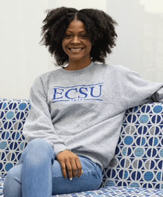 ECSU student wearing school shirt sitting on a coach smiling