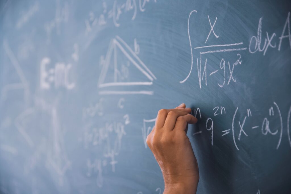 A hand writes mathematical equations in white chalk on a blue chalkboard.