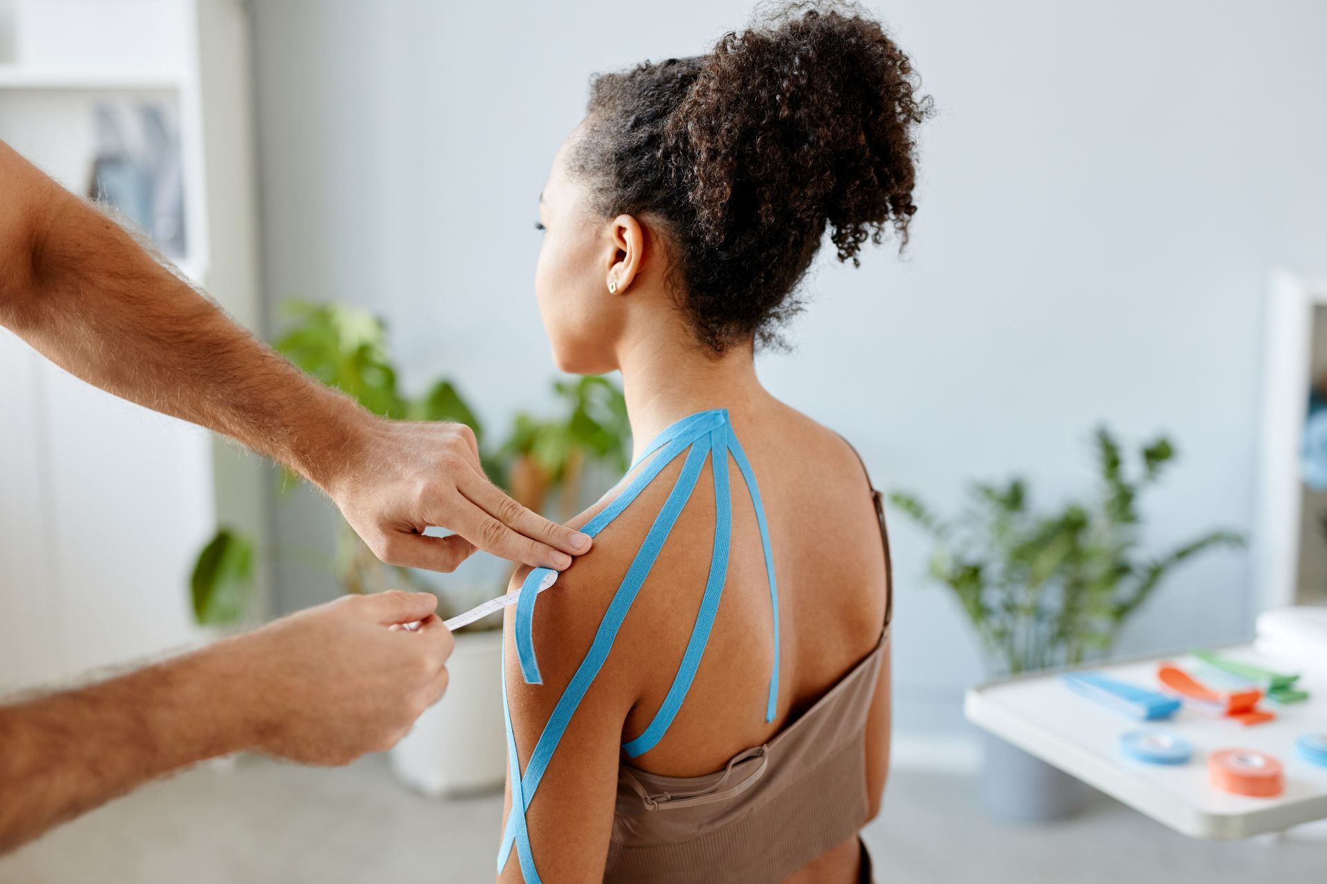 A woman receives kinesiology tape on her shoulder in a brightly lit room. The tape, light blue, is applied to her shoulder by a pair of hands.