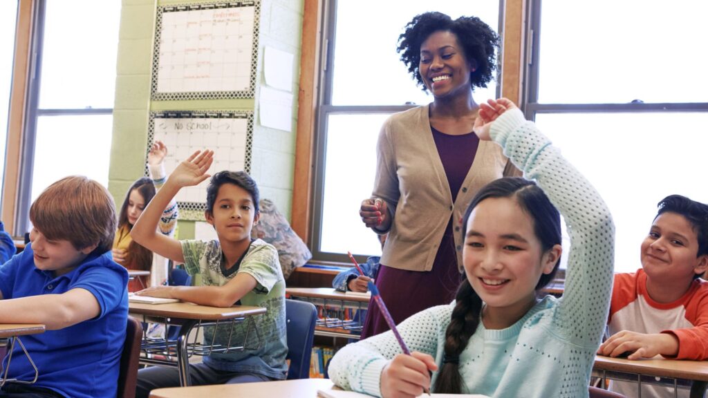 A teacher stands at the head of a classroom full of attentive students. Multiple children are raising their hands, hoping to answer the teacher's question.