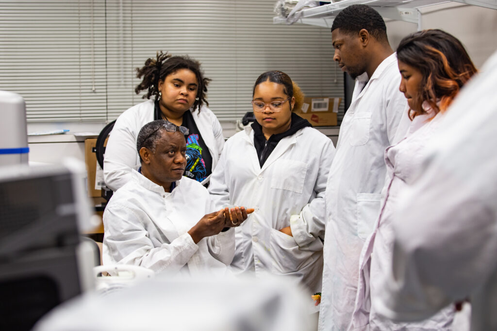 ECSU students gathered around their instructor for a lesson, all wearing lab coats.