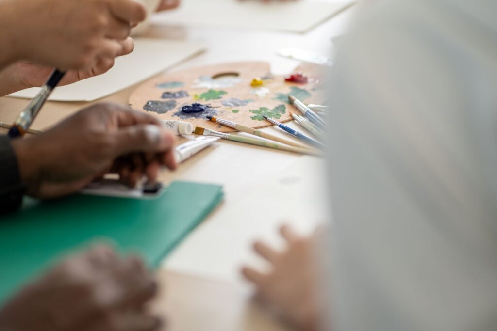 This bright photograph showcases a group of people engaging in the artistic activity of painting. The scene is set indoors, with the focus on hands holding paintbrushes and working on their canvases.