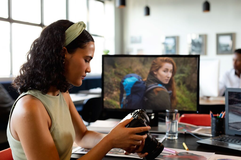 A woman with dark, wavy hair and a light-green top reviews photos on a digital camera at her office workspace, where a computer monitor displays an image of another woman in a rugged outdoor setting.