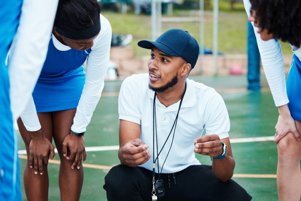 On a vibrant green and white sports field, an adult coach wearing a white polo shirt, black pants, and a dark cap kneels, gesturing with his hands while talking to a group of young adults.