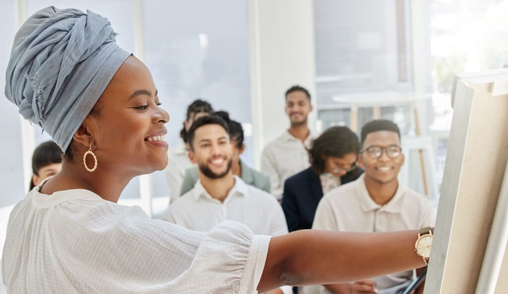 A smiling woman with a grey headwrap points to a white board in a bright office, where a group of colleagues are seated.