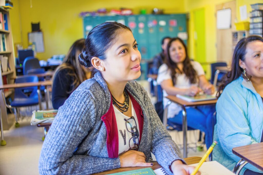 A teenaged student with hair pulled back sits at a desk in a brightly lit classroom.