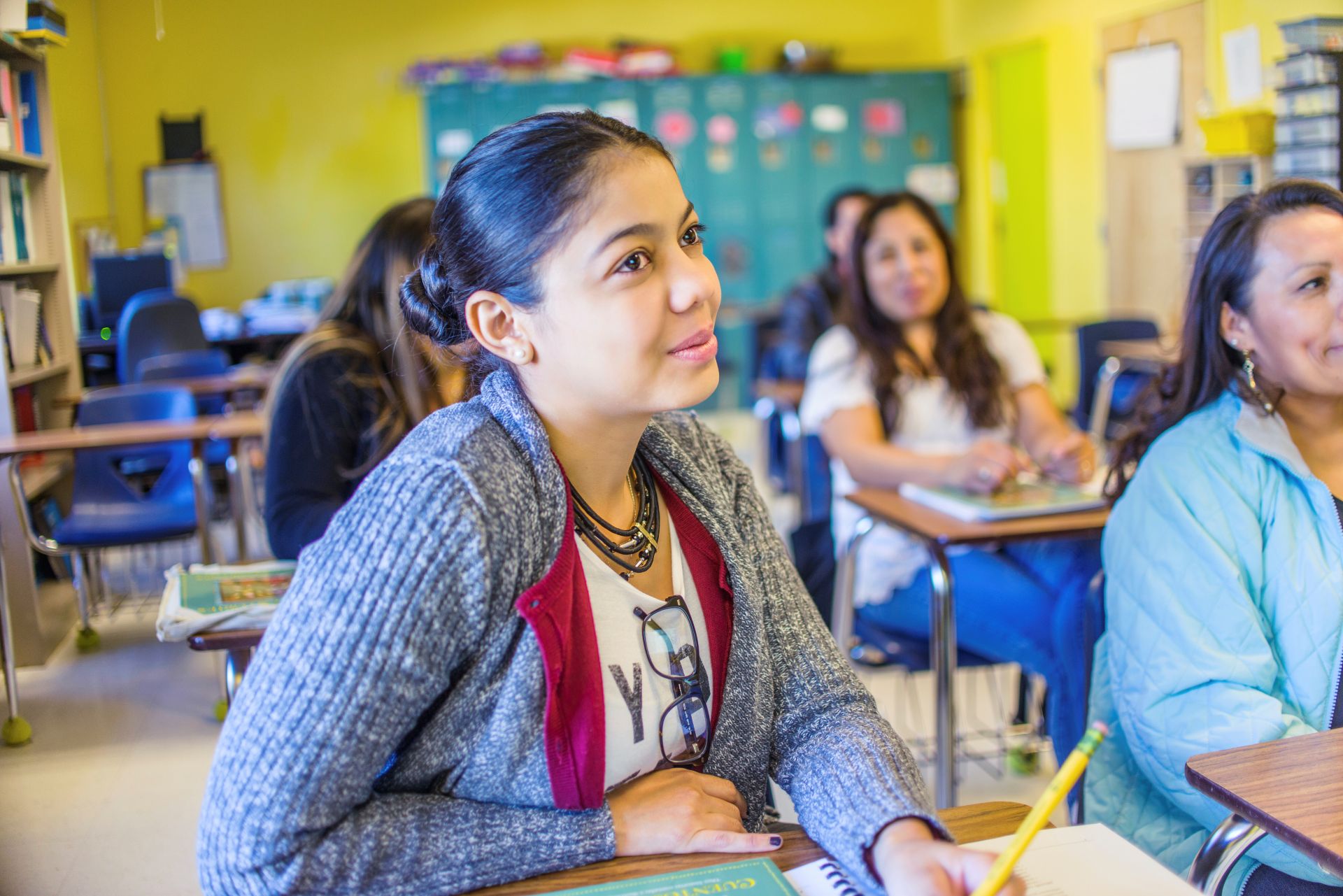 A teenaged student with hair pulled back sits at a desk in a brightly lit classroom.
