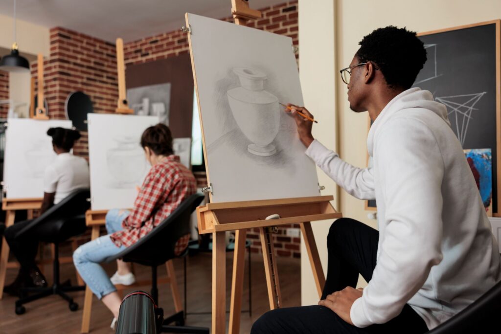 Three adults are attending an art class. One student is seated at the front and is using a pencil to draw the image of a vase onto a white canvas propped up on a wooden easel.