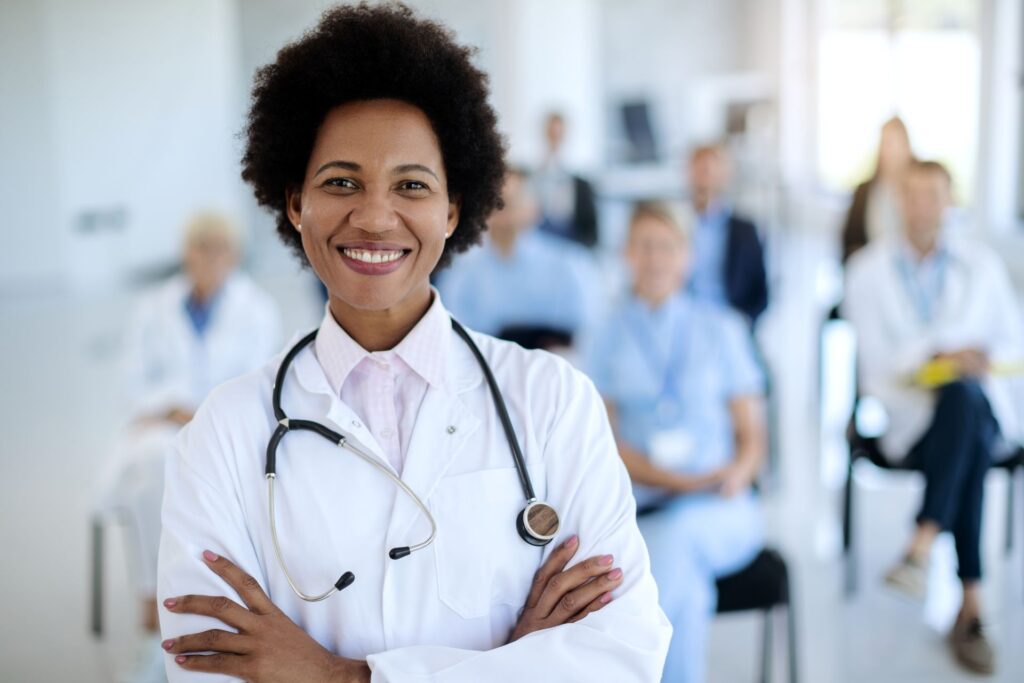 A smiling woman with dark curly hair wearing a white coat and stethoscope stands with her arms crossed, looking at the camera.