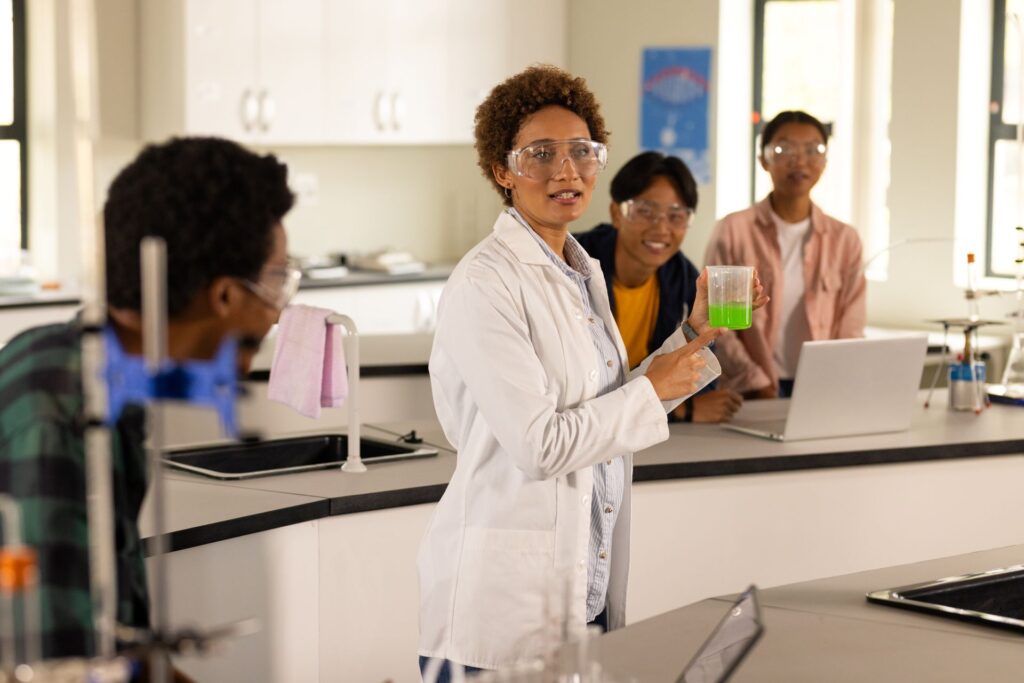 Students learn science in a bright laboratory setting. A woman with curly brown hair in a white lab coat and safety glasses holds a beaker with a green liquid.