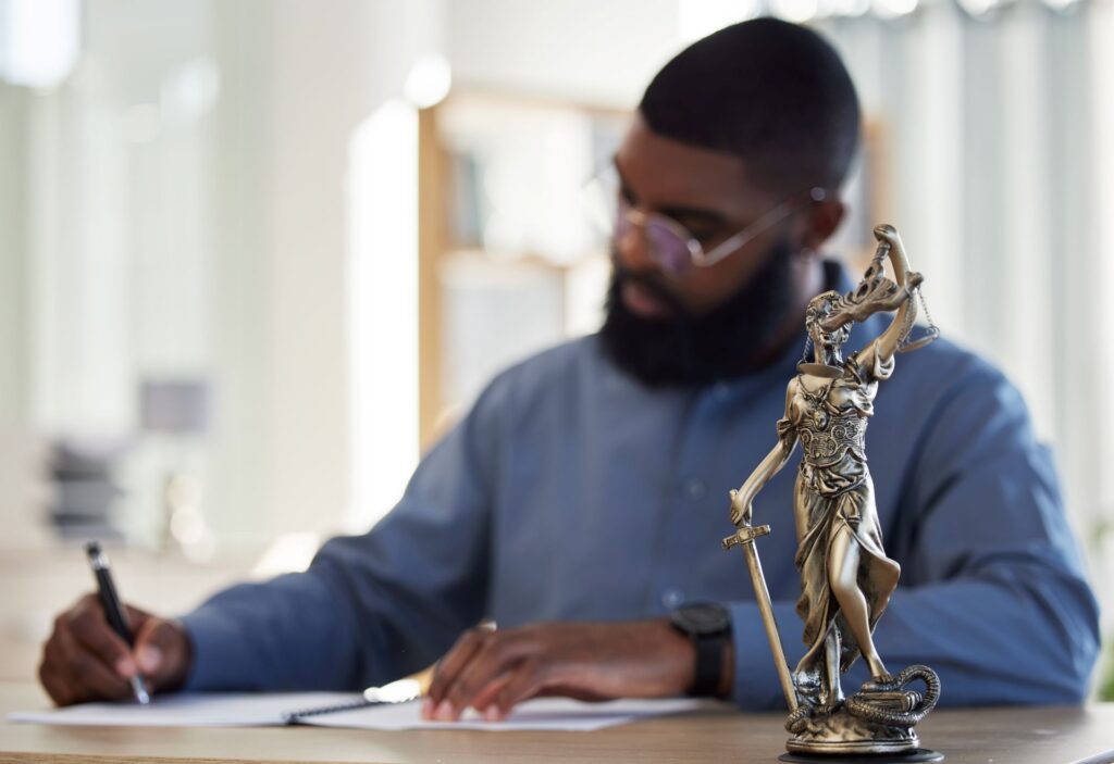 A man with short dark hair and a beard wearing glasses is writing on paper in an office.