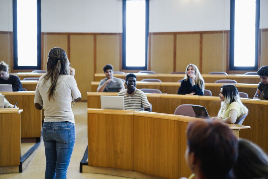 A woman with long brown hair, wearing jeans and a beige long-sleeved shirt, stands facing a classroom of students.