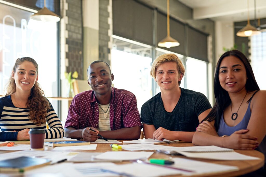 Four smiling young adults are seated at a table in a brightly lit, urban workplace setting.