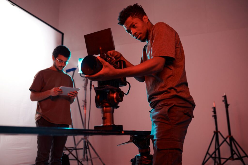 Two men are inside a studio space. One with curly hair and glasses holds a tablet, and the other adjusts a large video camera.
