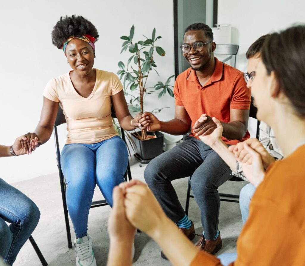 A group of smiling adults sit in a circle holding hands indoors.
