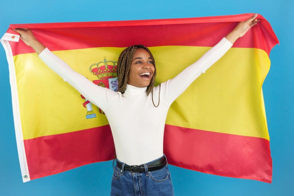 A young woman smiles while holding a Spanish flag against a light blue background. 