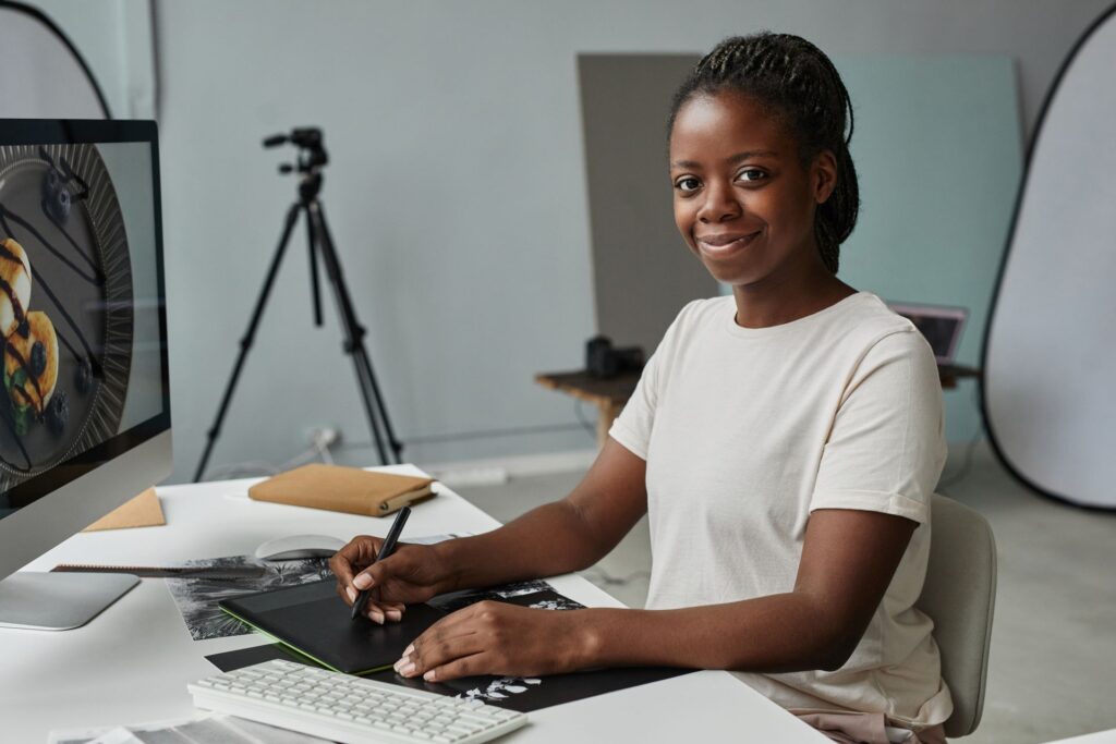 In a studio setting, a smiling woman with braided dark hair sits at a white desk and works on a graphics tablet, her hand holding a stylus.