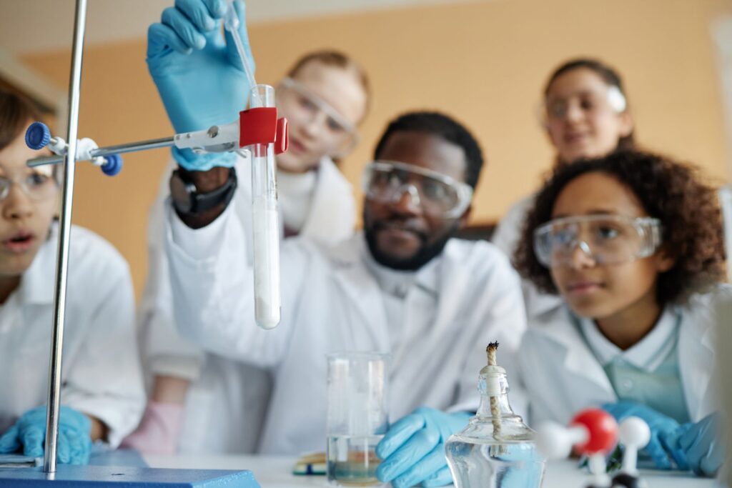 A small group gathers around a laboratory bench as an adult with short dark hair wearing a white coat and safety glasses performs an experiment.