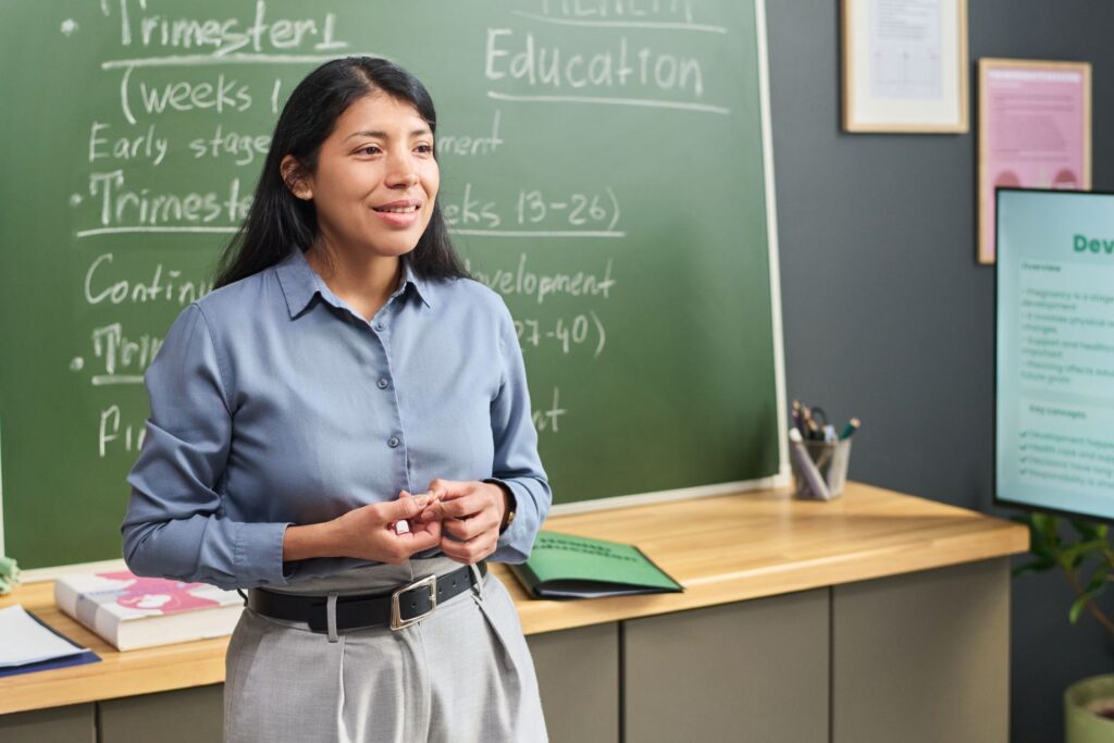 A woman stands in front of a green chalkboard with handwritten notes about health education.