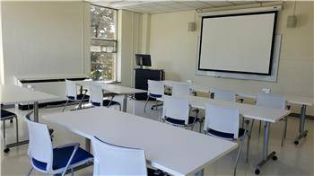 Full room shot of room 214 in the library. An instruction room with tables and chairs with an area for a speaker and presentation.