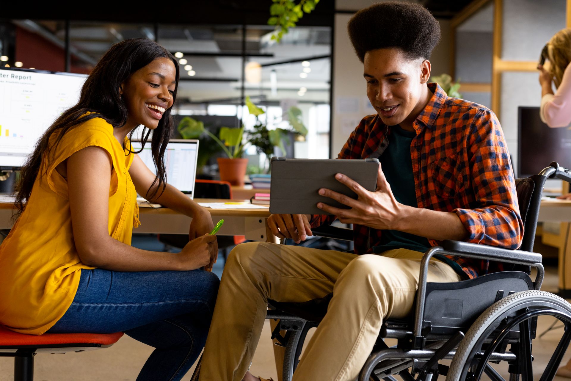 In a bright and modern office space, a young woman with long dark hair wearing a yellow top and jeans sits next to a young man using a tablet while seated in a wheelchair.