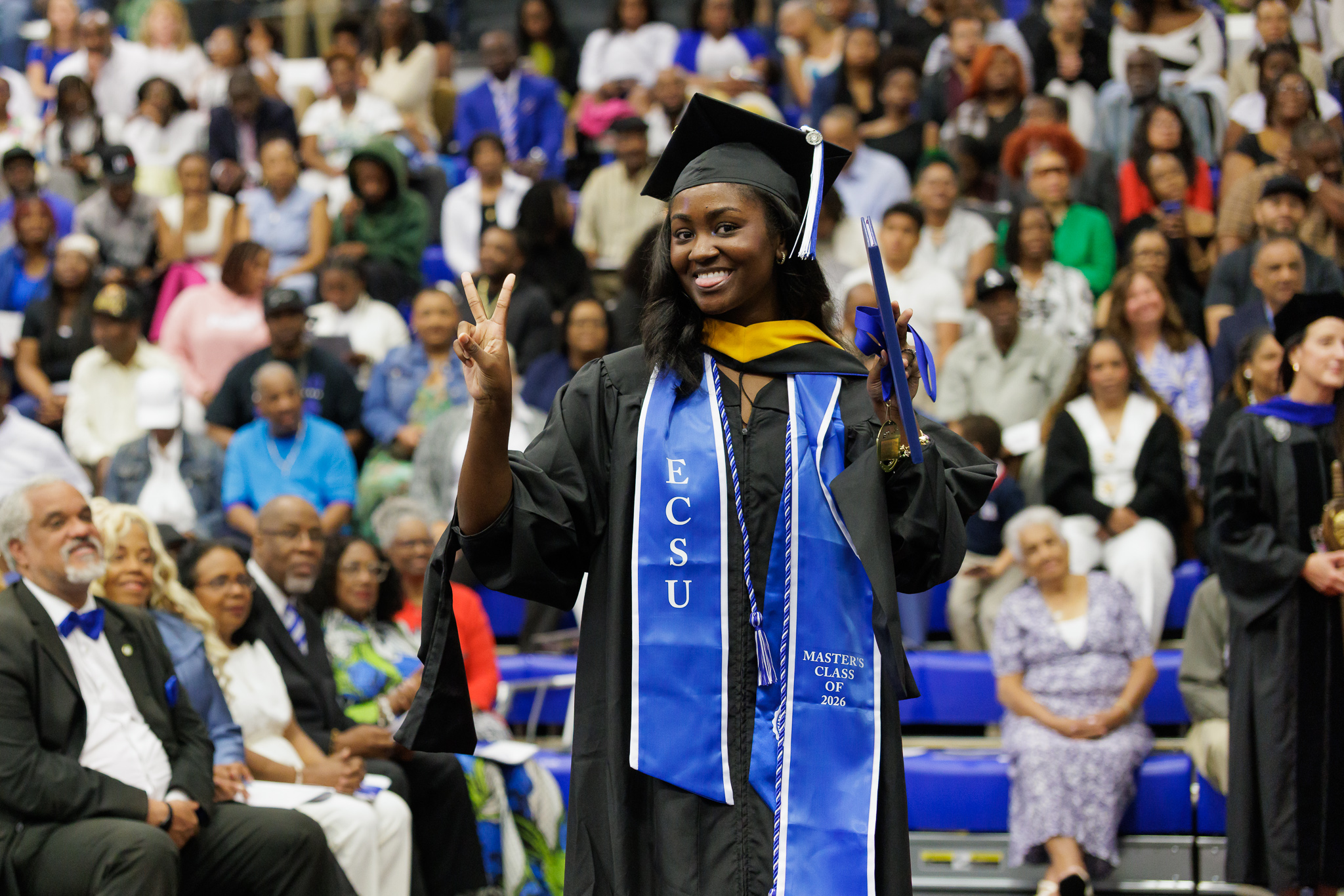 Graduate in a black cap and gown stands in front of a crowded auditorium, holding a diploma and making a peace sign. A blue stole with “ECSU” and “Master’s Class of 2026” hangs around the graduate’s neck, while an audience fills the background seats.
