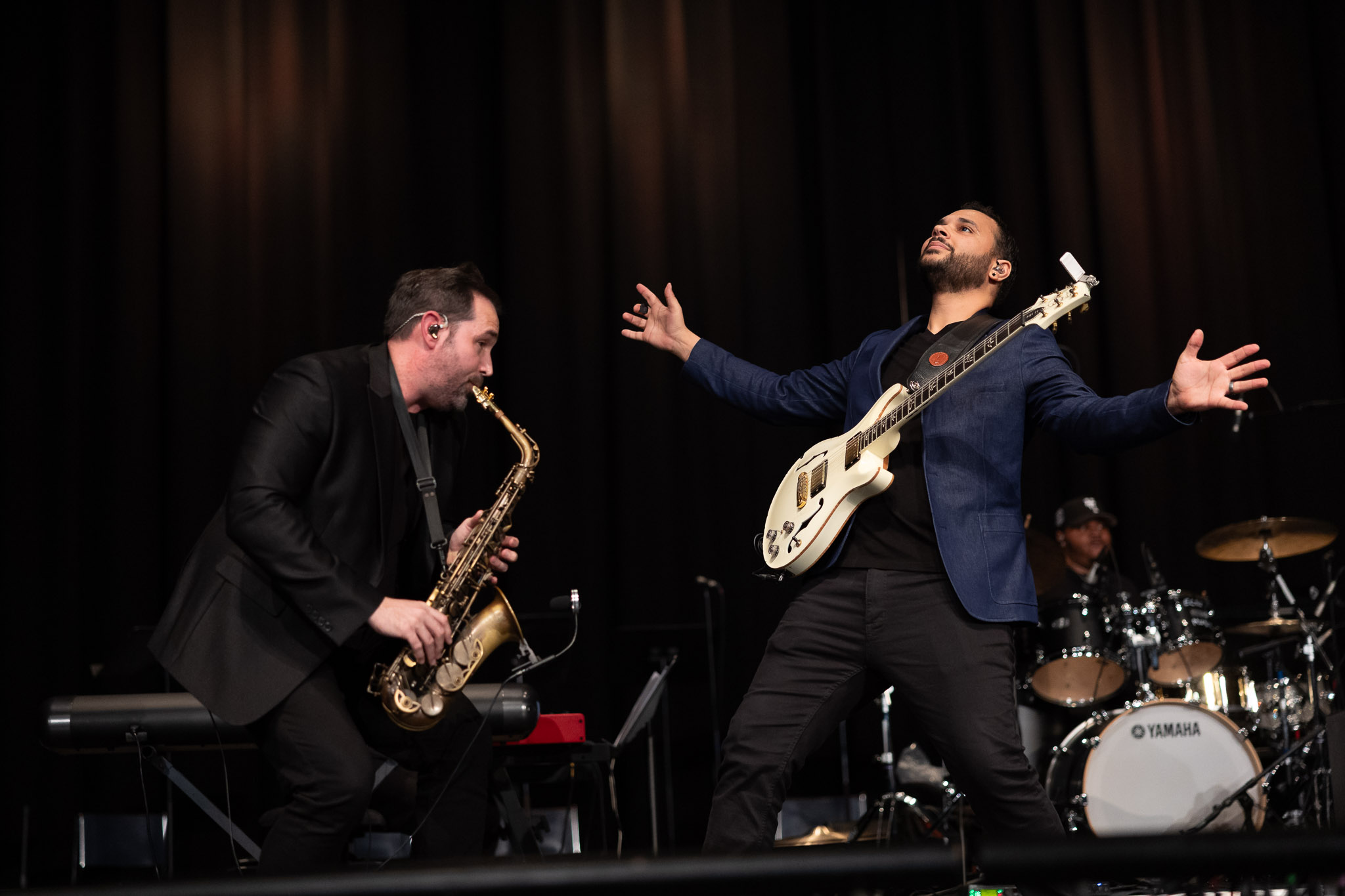 Two musicians perform on a dimly lit stage. One musician plays a saxophone while leaning forward, and another stands with an electric guitar, arms extended outward. A drum set and additional band equipment are visible in the background under stage lighting.