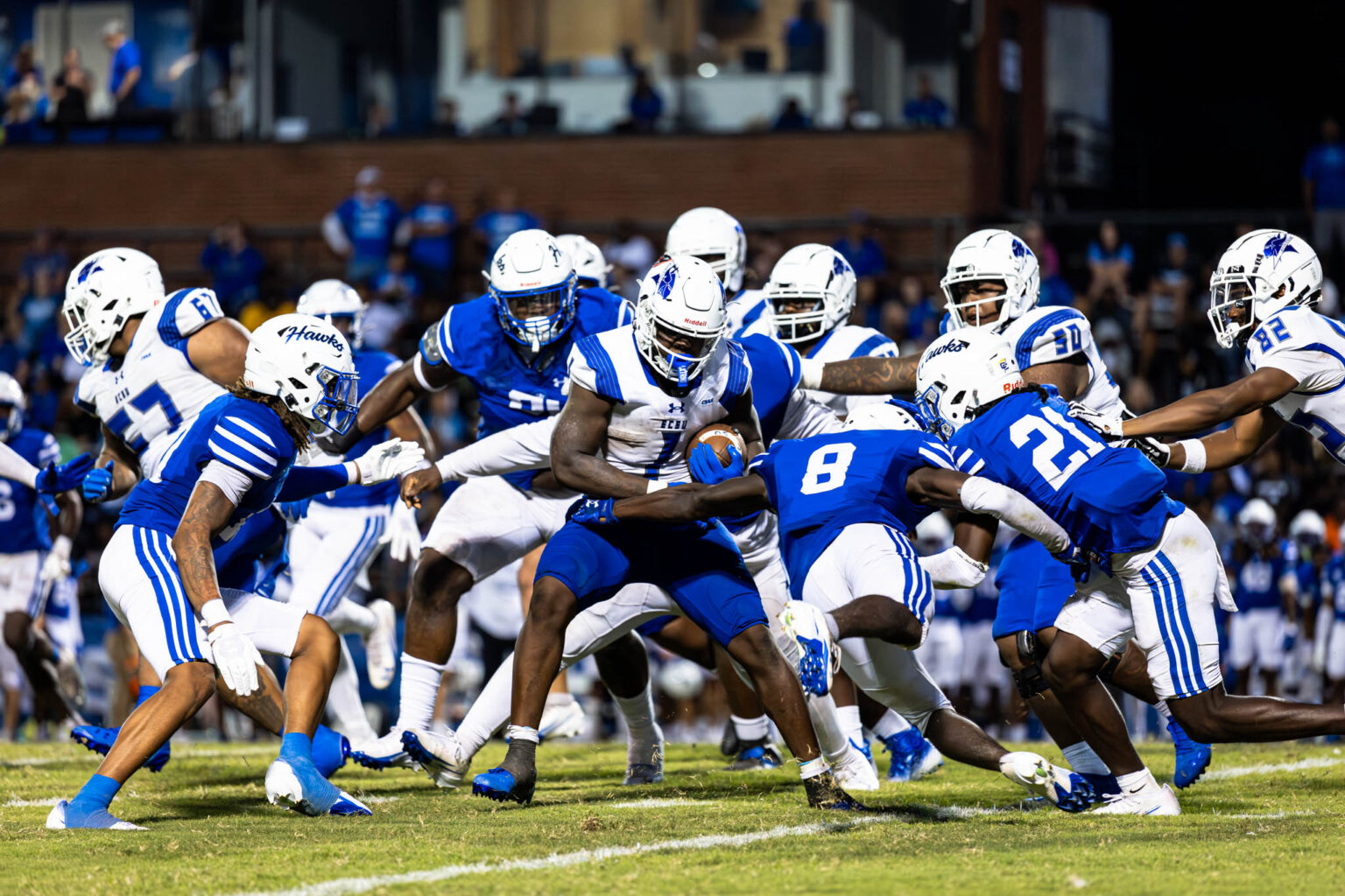 A football player in a white jersey carries the ball while multiple opposing players in blue uniforms converge to tackle him during a game. Players from both teams cluster around the runner on a grassy field with spectators in the background.
