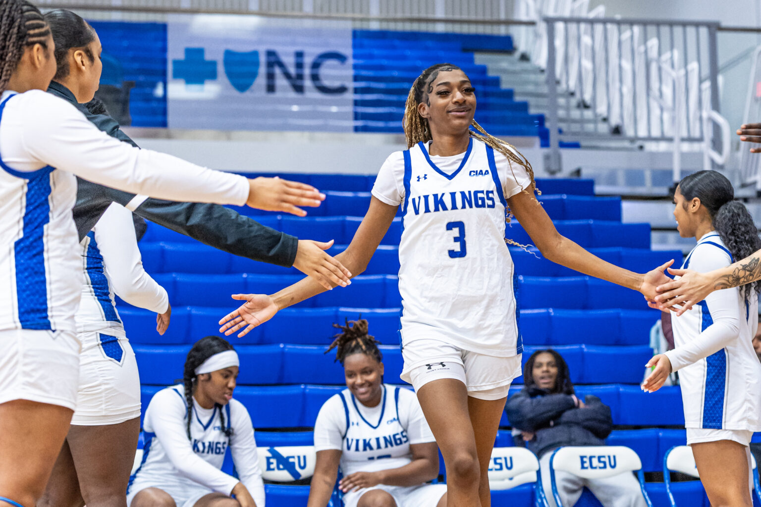ECSU women’s basketball player wearing a white “Vikings” uniform with the number 3 walks between teammates, exchanging high-fives as she is introduced on the court, with blue bleachers in the background.