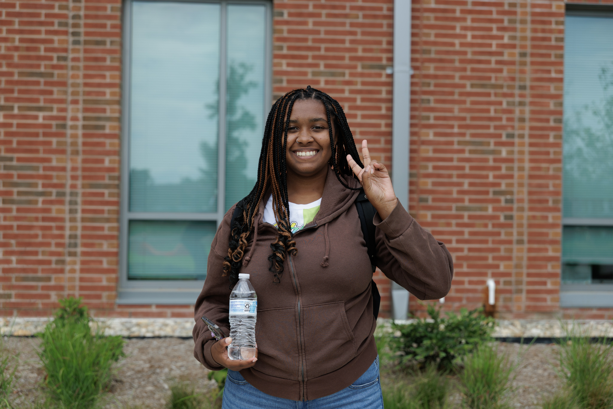 Student stands outdoors in front of a brick campus building, smiling at the camera while holding a water bottle and making a peace sign. The student wears a brown hoodie and carries a backpack, with greenery and windows visible in the background.