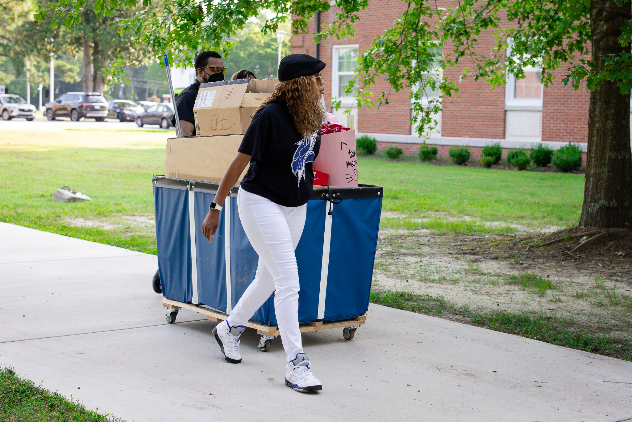 Two people move a cart loaded with boxes and belongings along a campus walkway, likely during move-in. One person walks in front while another pushes the cart, with trees and a brick campus building in the background.