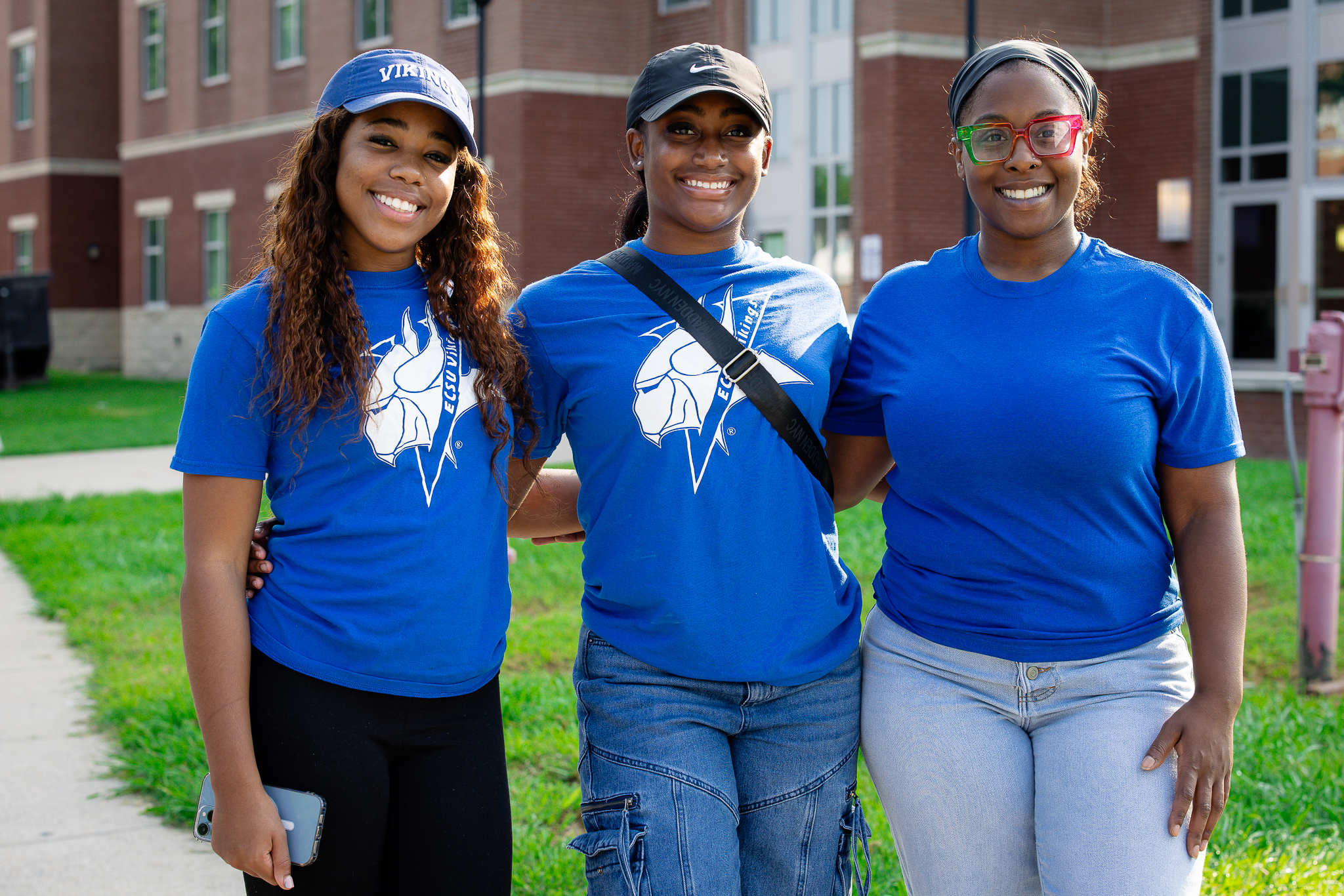 Three ECSU students stand outdoors in front of a residence hall, smiling with arms around each other while wearing matching blue T‑shirts with the Viking logo.