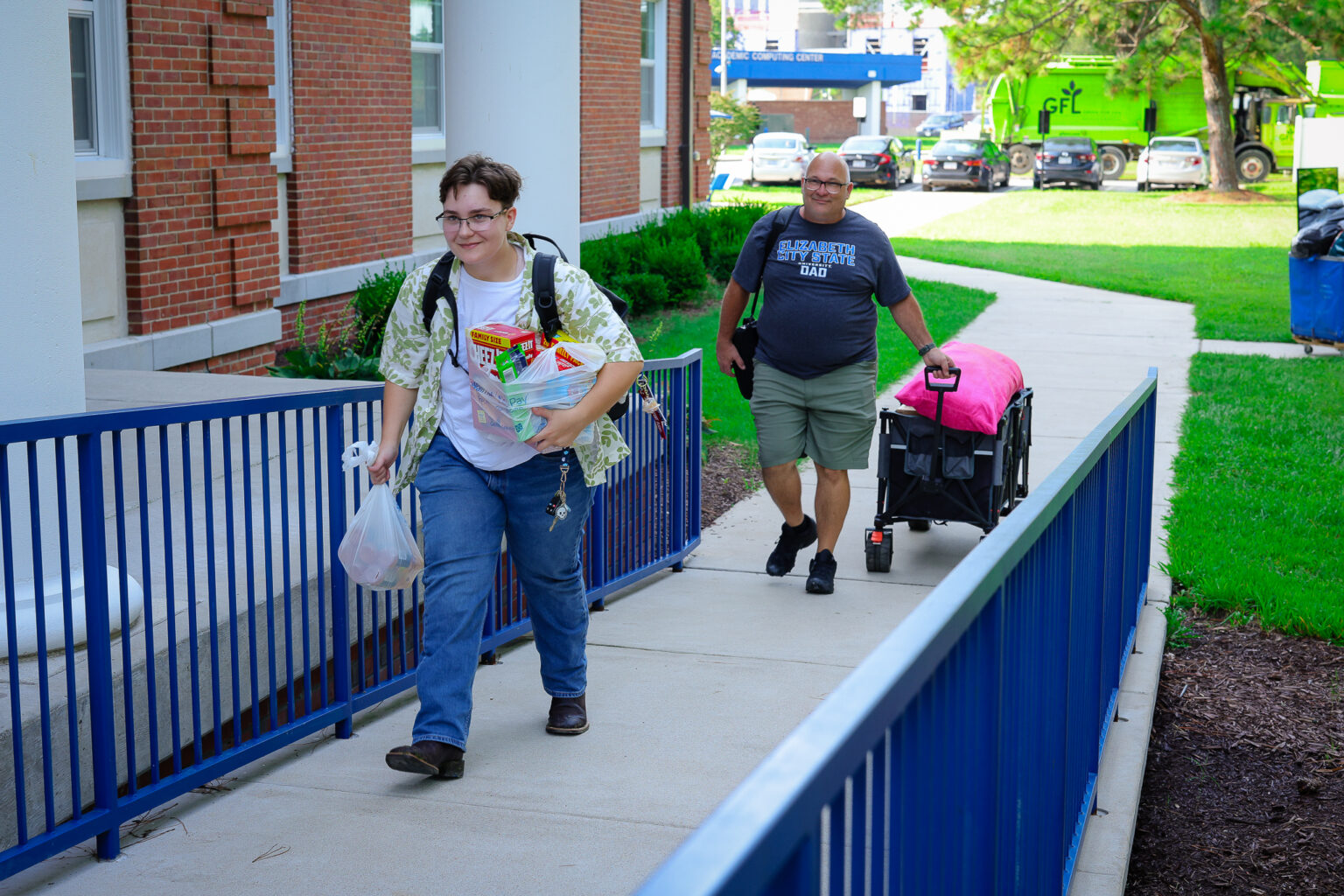 Student carries bags and groceries up a campus walkway toward a residence hall while a family member pulls a wagon loaded with belongings behind them during move-in.
