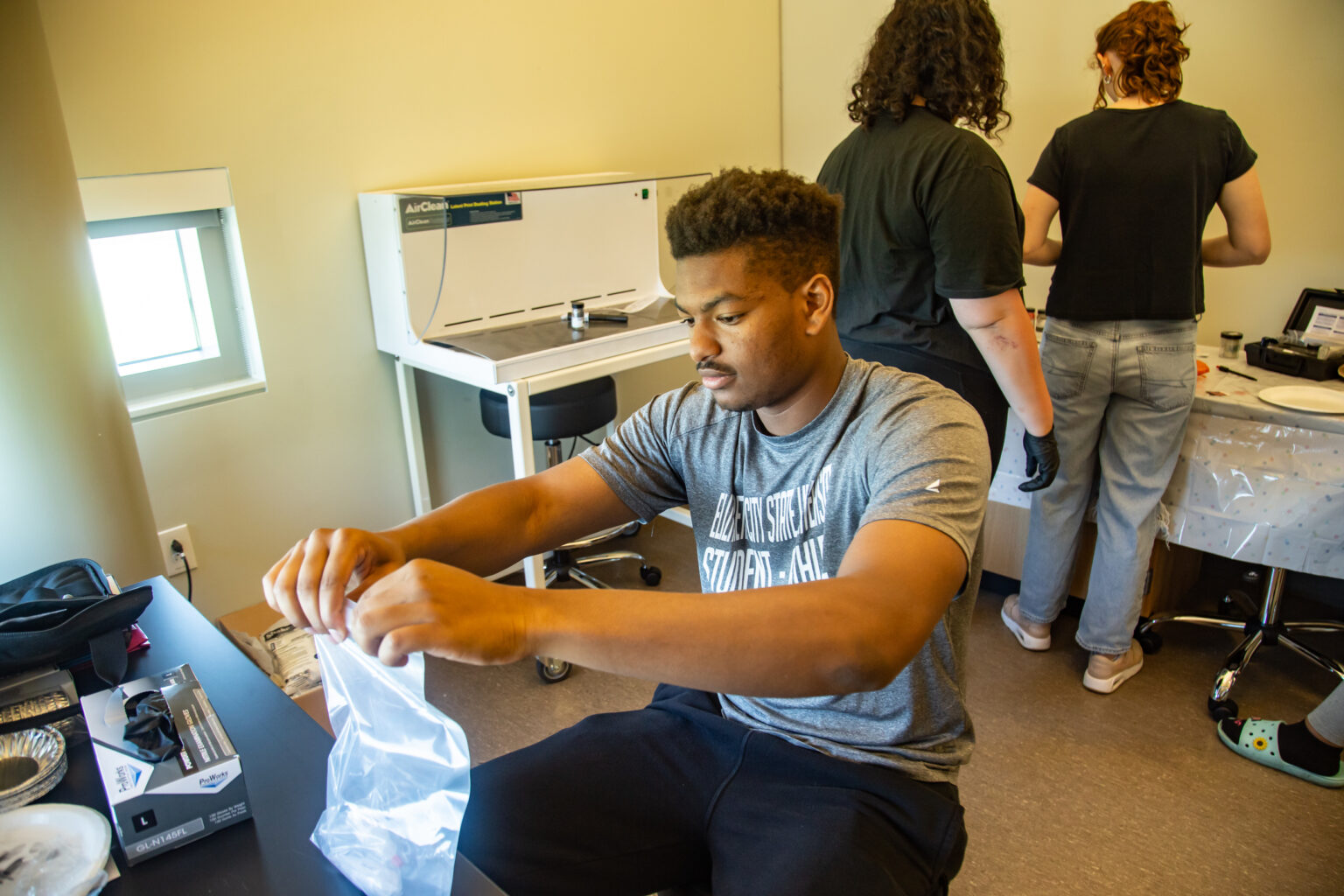 Student sits at a lab table placing materials into a plastic bag while two others work at a counter in the background, wearing gloves and handling supplies in a forensic or science lab setting.